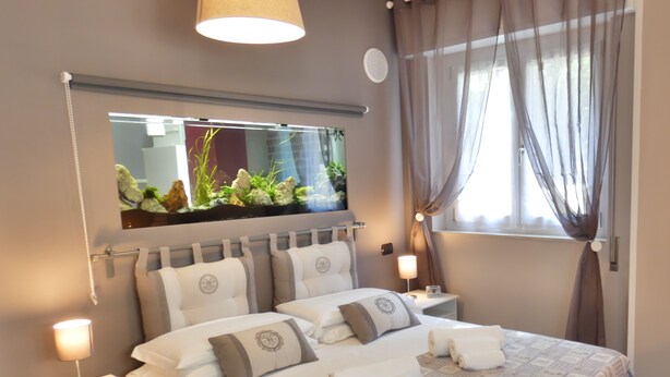 A neatly made bed with a patterned bedspread, two bedside tables with lamps, a large mirror above the bed, and a window with sheer curtains. - Charming House in Old Town Desenzano (Desenzano del Garda)