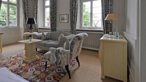 A well-lit room with a sofa, armchair, and a small table with a lamp. There are windows with curtains, a radiator, and a patterned rug on the floor. - Hotel Herrenhaus von Löw (Bad Nauheim)