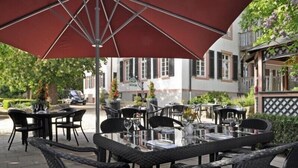 An outdoor dining area with tables and chairs under a large umbrella, surrounded by greenery and a building with a lattice facade. - Hotel Herrenhaus von Löw (Bad Nauheim)