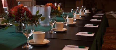 A conference table set with white cups, saucers, and wine glasses, with a green tablecloth and floral centerpieces.
