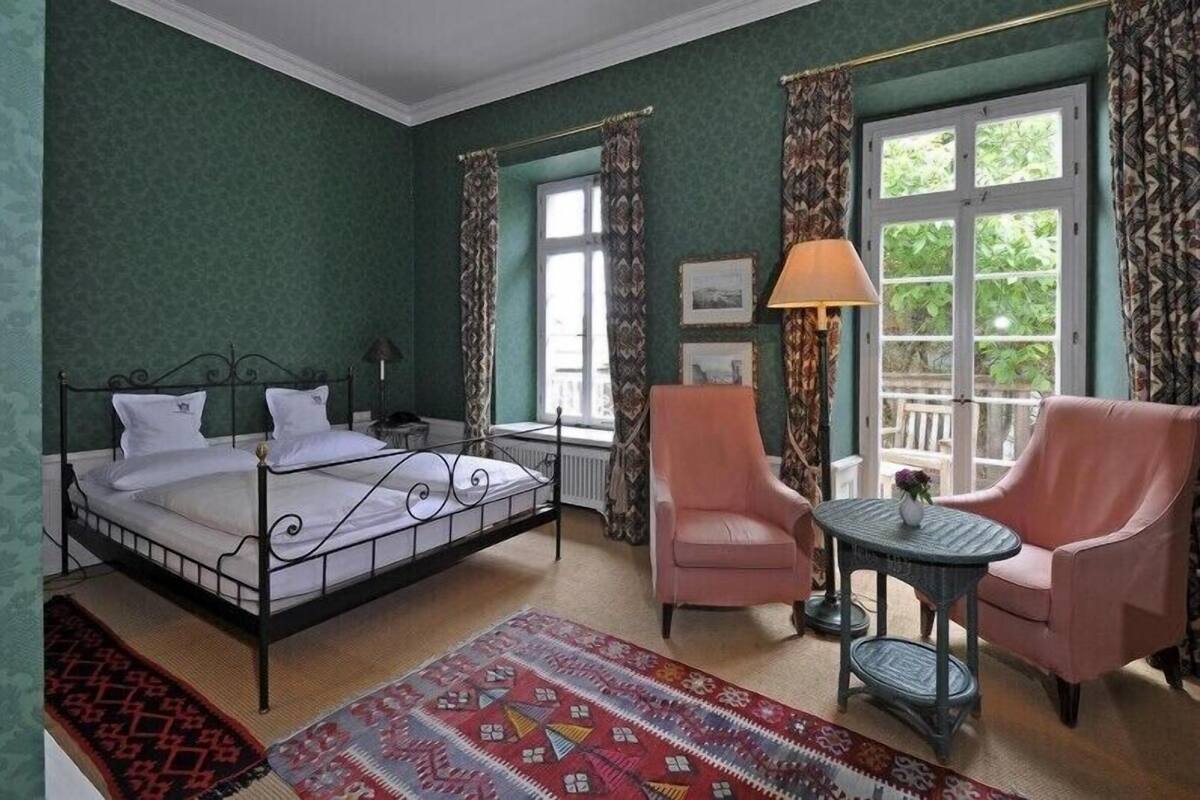 A bedroom with a four-poster bed, two chairs, a small table, and a window with curtains.