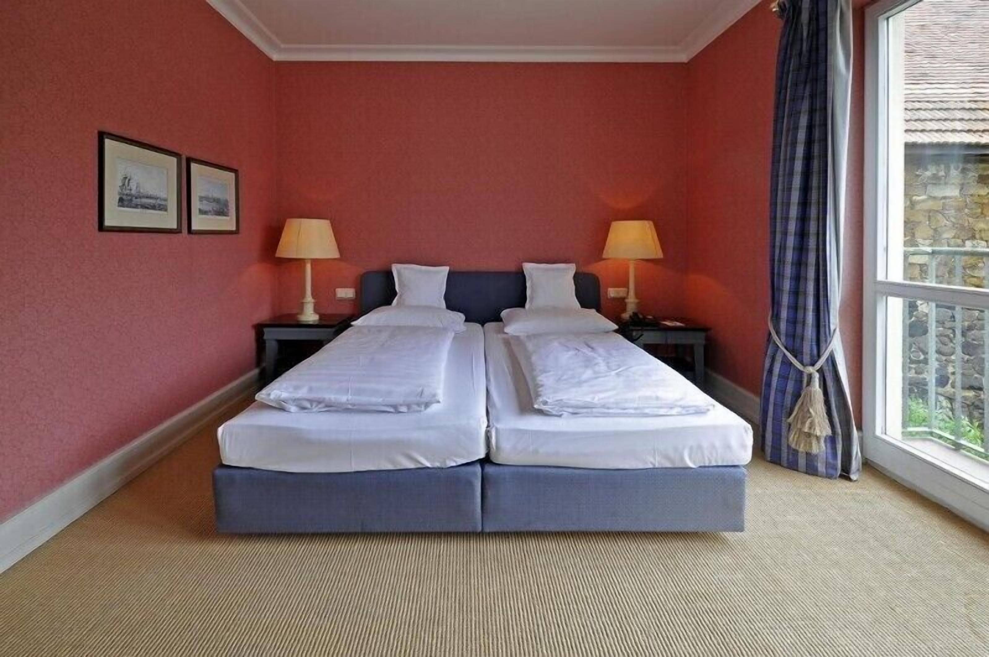 A double bed with two pillows, a nightstand with a lamp, and a window with a view of a stone building.