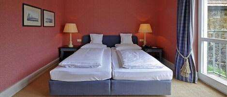 A double bed with two pillows, a nightstand with a lamp, and a window with a view of a stone building.