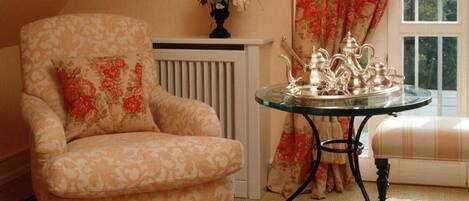 A cozy room with a floral-patterned armchair, a small table with a tea set, and a window with a floral curtain.