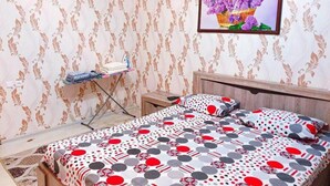 Ein Schlafzimmer mit strukturiertem Wandpapier, einem Bett mit gepunktetem und geometrischem Bettbezug, einem Nachttisch, einem an der Wand hängenden Bild und einem kleinen Bügelbrett mit Kleidung.