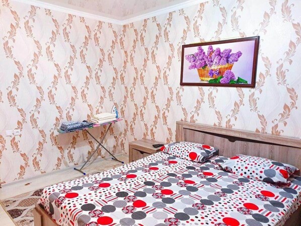 Ein Schlafzimmer mit gemustertem Tapetenbelag, einem Bett mit Kissenüberwurf in Punkte und Streifen, einem kleinen Tisch mit gefalteter Kleidung und einem gerahmten Bild an der Wand.