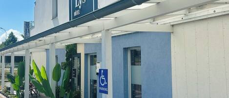 Um hotel chamado "Le Lys" com área de estar coberta, sinal de estacionamento para deficientes e plantas em vasos ao longo da calçada.