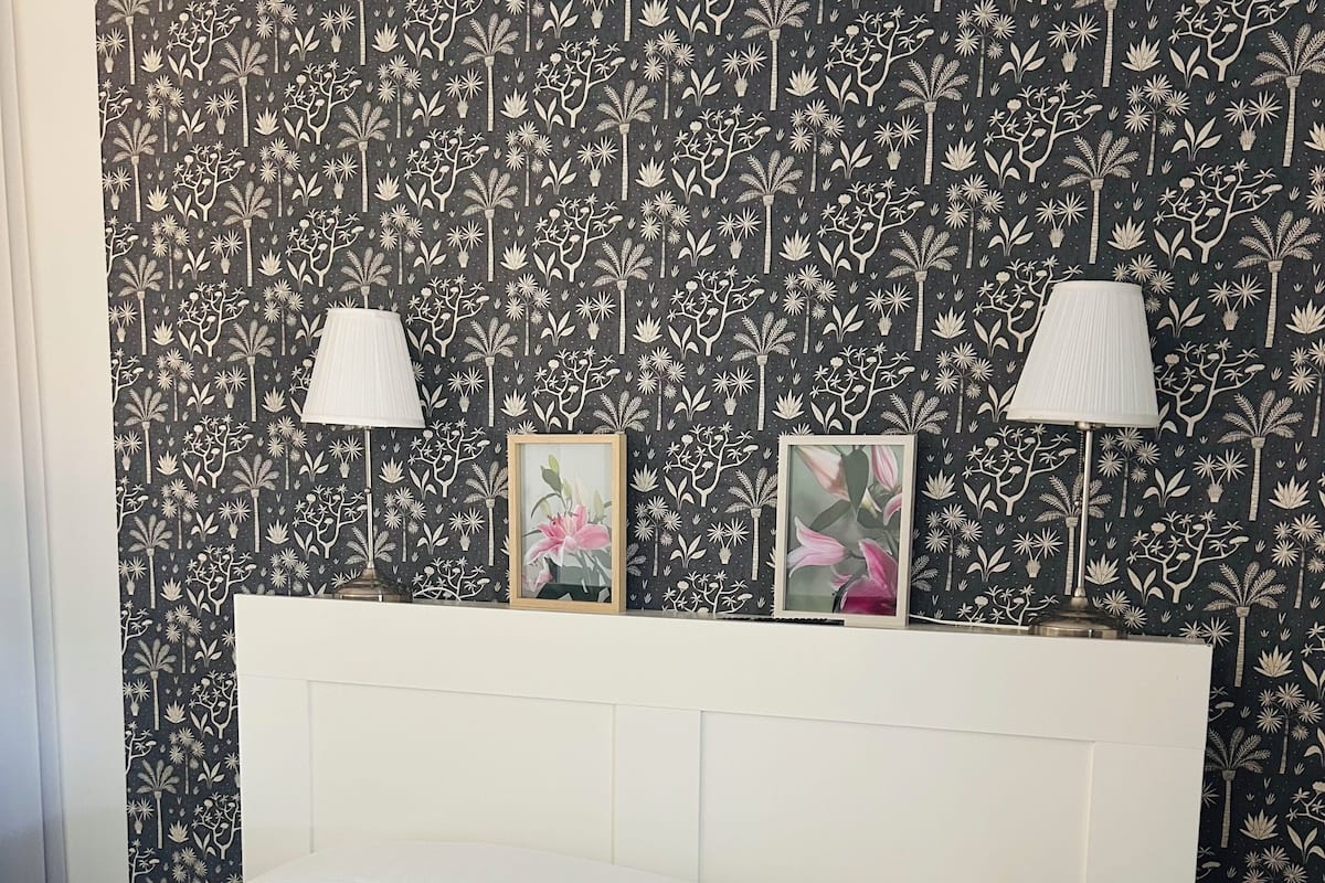 Une chambre à coucher avec un lit blanc, une tête de lit agrémentée de deux lampes fixées au mur, et un papier peint à motifs floraux.