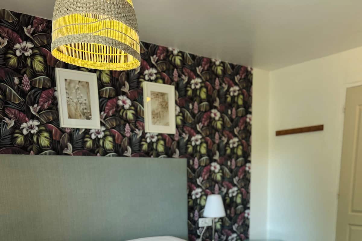Un lit bien fait dans une chambre d’hôtel, avec un papier peint à motifs floraux, un luminaire suspendu et une table de chevet avec une lampe.
