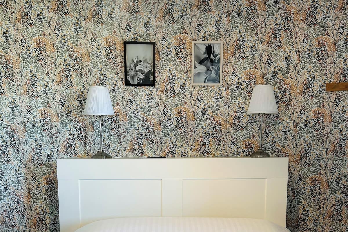 Une chambre à coucher avec un papier peint à motifs, un lit blanc, deux lampes de chevet et un luminaire suspendu.