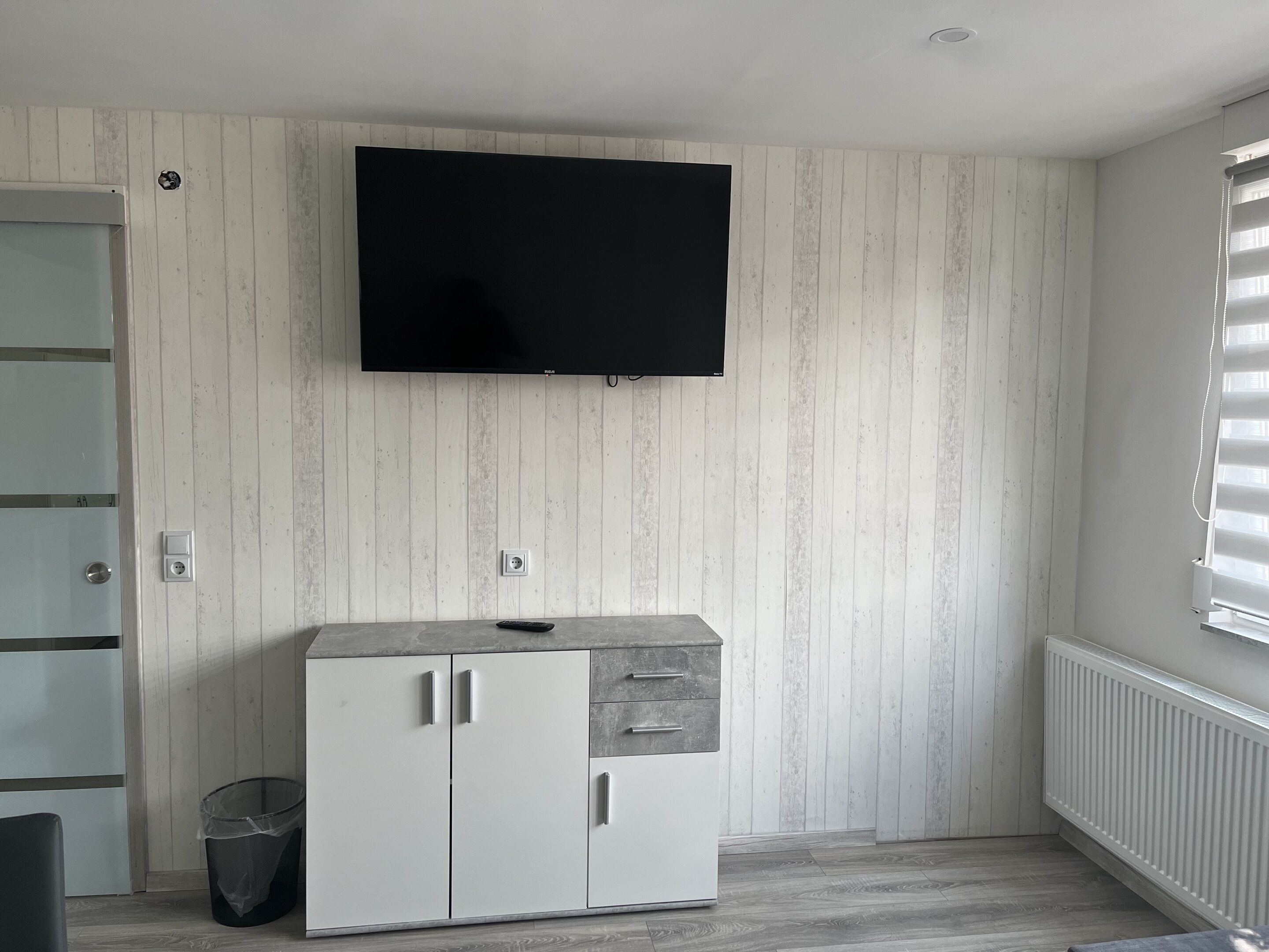 Una habitación con televisor incorporado, armarios blancos, un cesto de basura y una ventana con persianas.