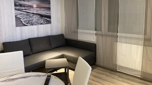 Un salon moderne avec un canapé d’angle gris, une petite table ronde sur laquelle se trouve une télécommande, et une grande fenêtre ornée de rideaux légers.