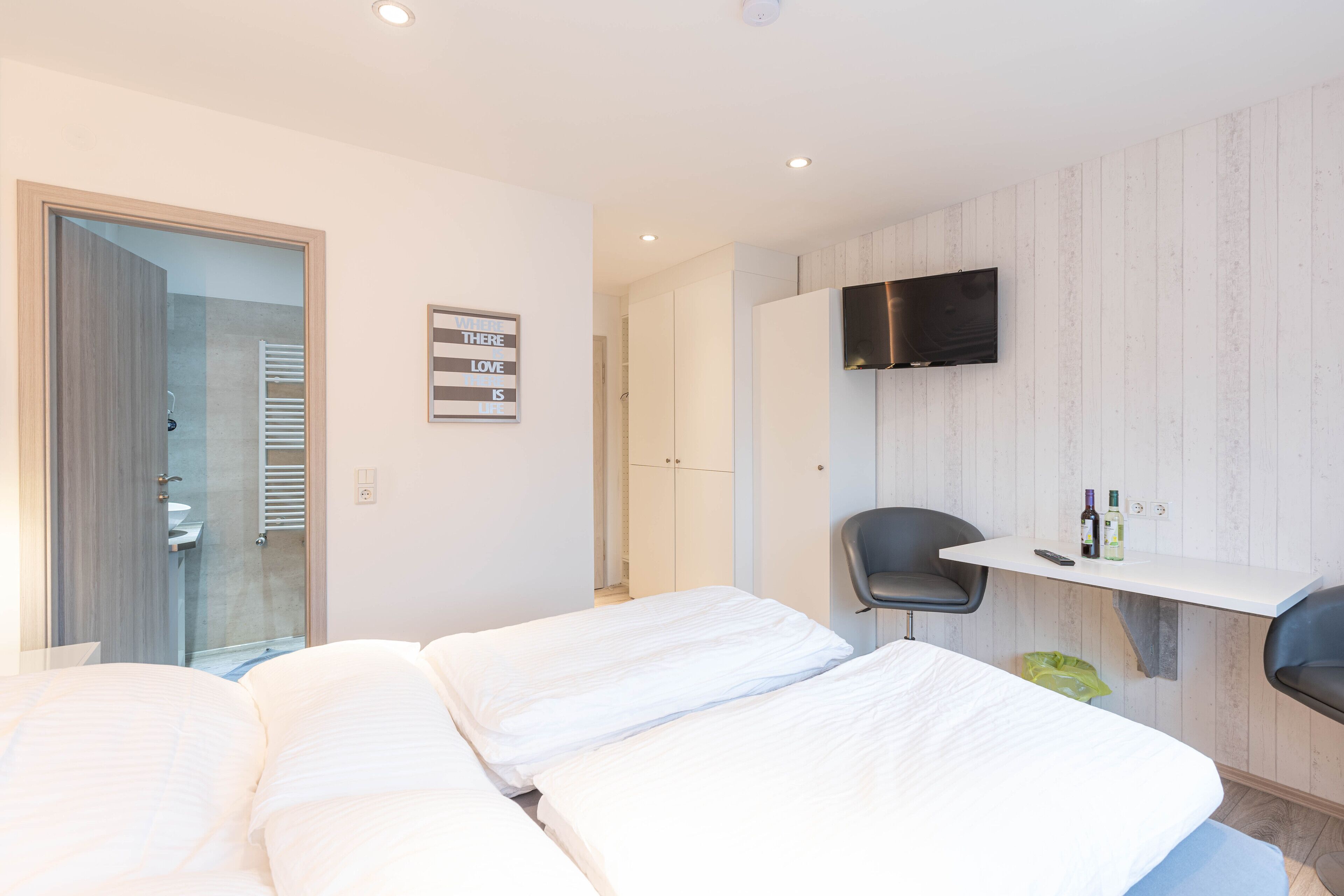 Una habitación de hotel moderna con dos camas, un televisor montado en la pared, un escritorio pequeño con una silla y un baño visible a través de una puerta de vidrio.