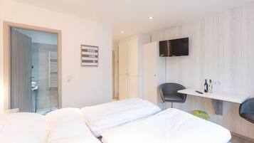 Una habitación de hotel moderna con dos camas, un televisor montado en la pared, un escritorio pequeño con una silla y un baño visible a través de una puerta de vidrio.