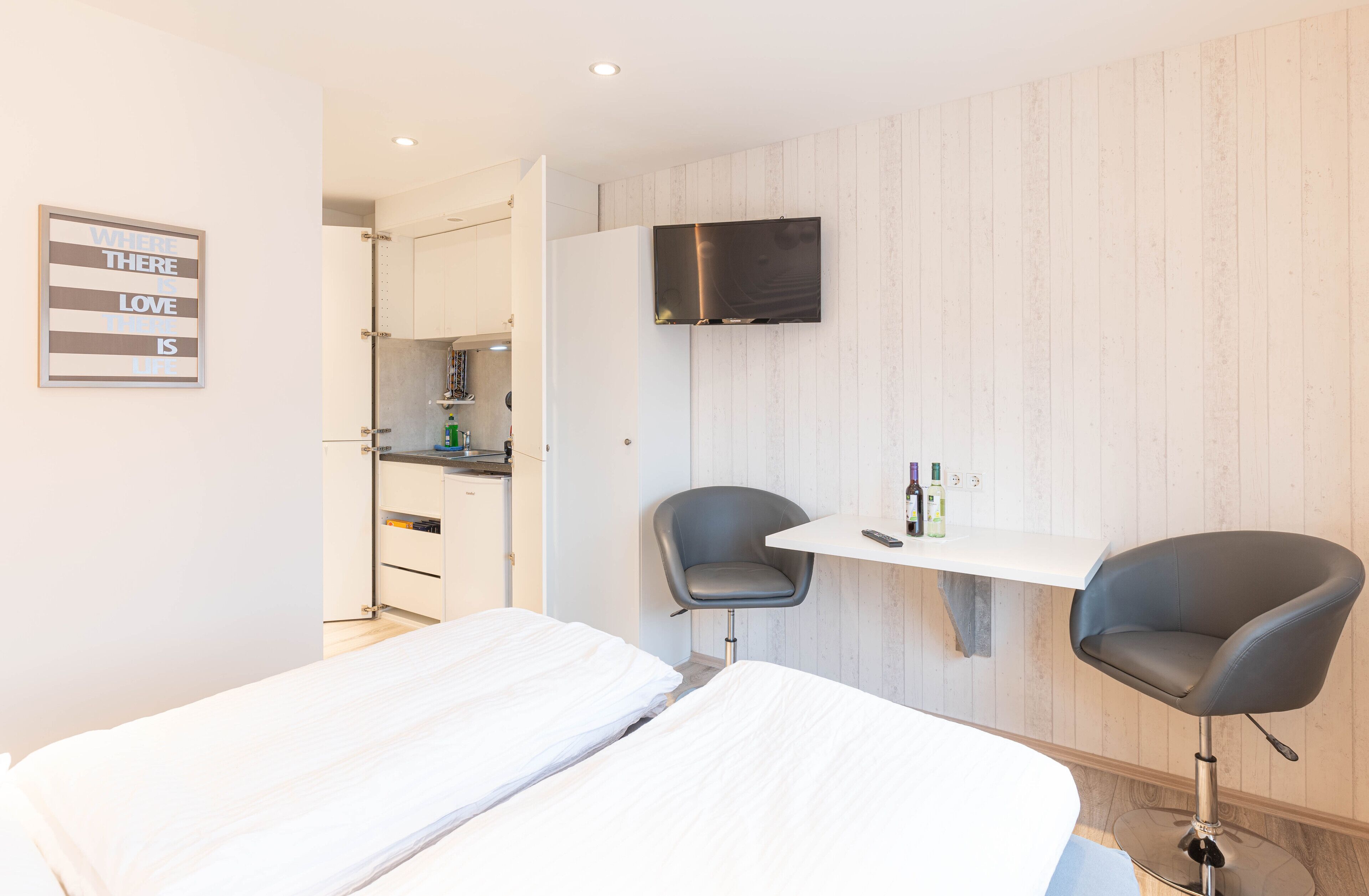 A hotel room with two beds, a small desk with chairs, a TV, and a kitchen area visible through an open door.