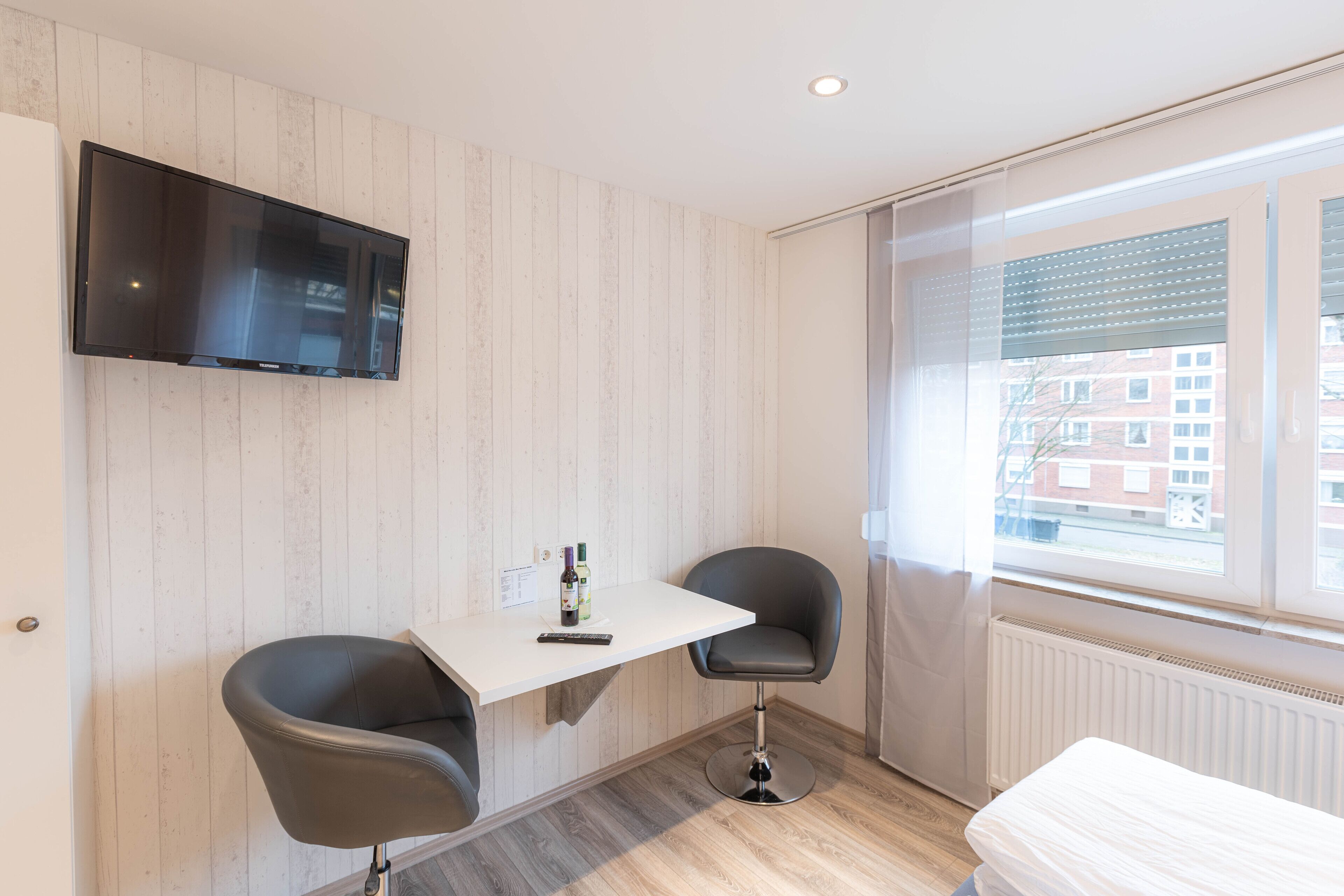 Una habitación moderna con un televisor de pantalla plana montado en la pared, una pequeña mesa con dos sillas, una cama y una ventana con persianas.