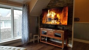 Een woonkamer met een open haard, een televisie erboven, een houten kastje eronder en een raam met jaloezieën.