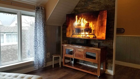 A living room with a fireplace, a television mounted above it, a wooden cabinet below, and a window with blinds.
