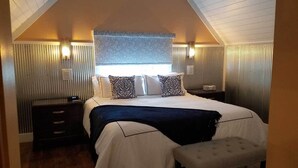 Een slaapkamer met een schuine plafond, een groot bed met gestreepte kussens, nachtlampen en een bankje.