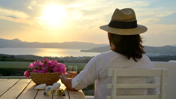 Una persona sentada a una mesa de madera con una cesta de flores, un vaso de bebida y un par de gafas de sol. La mesa está al aire libre, con vistas a un cuerpo de agua y montañas a lo lejos.