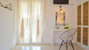 Una habitación moderna con un ventanal grande, un televisor de pantalla plana montado en la pared, una pequeña mesita consola con objetos decorativos y una silla original de alambre.