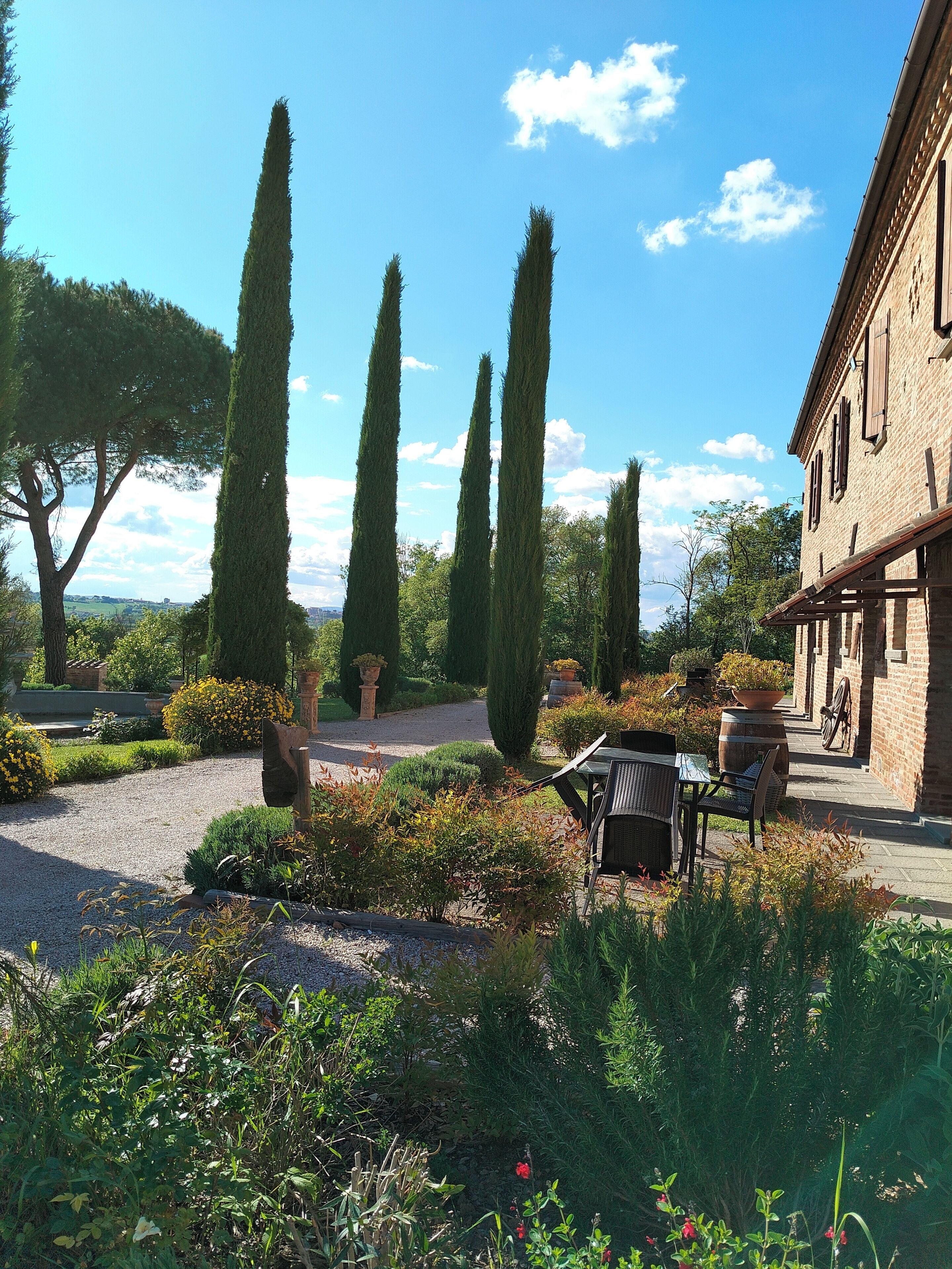 Un jardin avec un chemin de gravier, de grands cyprès et un bâtiment en briques doté d’un patio couvert.