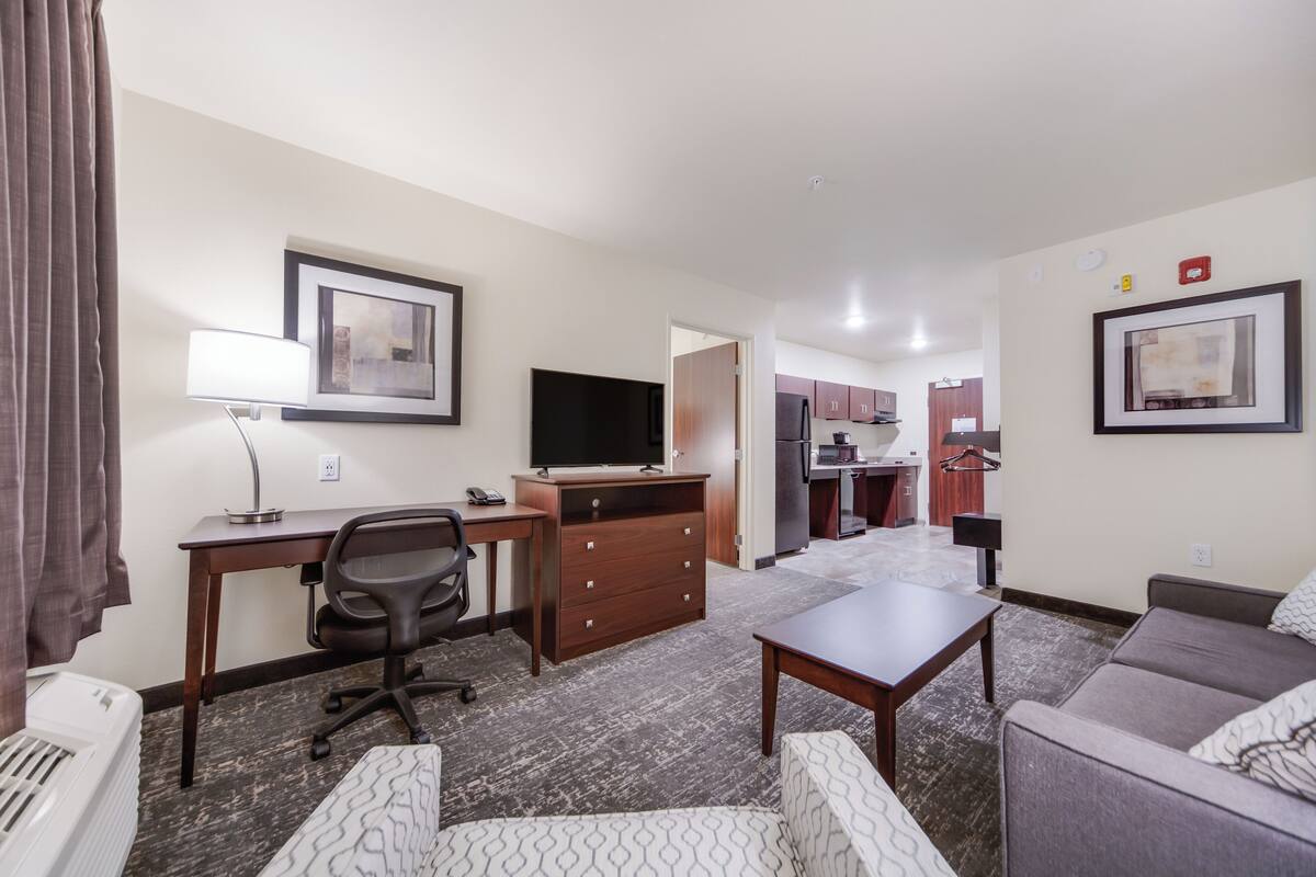 A hotel room with a desk, chair, sofa, and a kitchenette in the background.