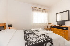 Ein Schlafzimmer mit einem Bett, einer Holzkommode, einem Sessel, einem Schreibtisch, einer Lampe und einem Fenster mit Vorhängen.