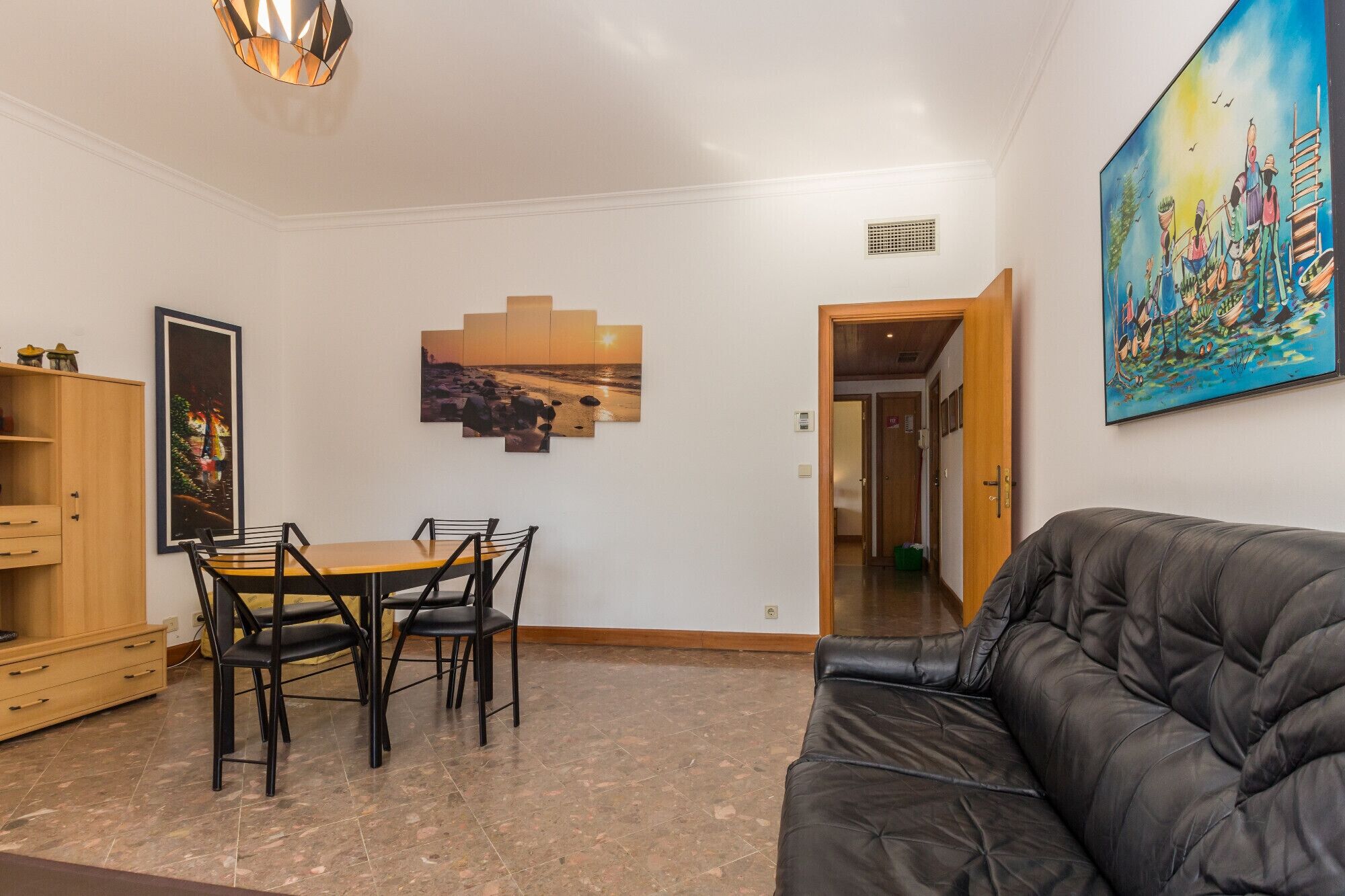 A living room with a black leather sofa, a dining table with chairs, a wooden cabinet, and a painting on the wall.