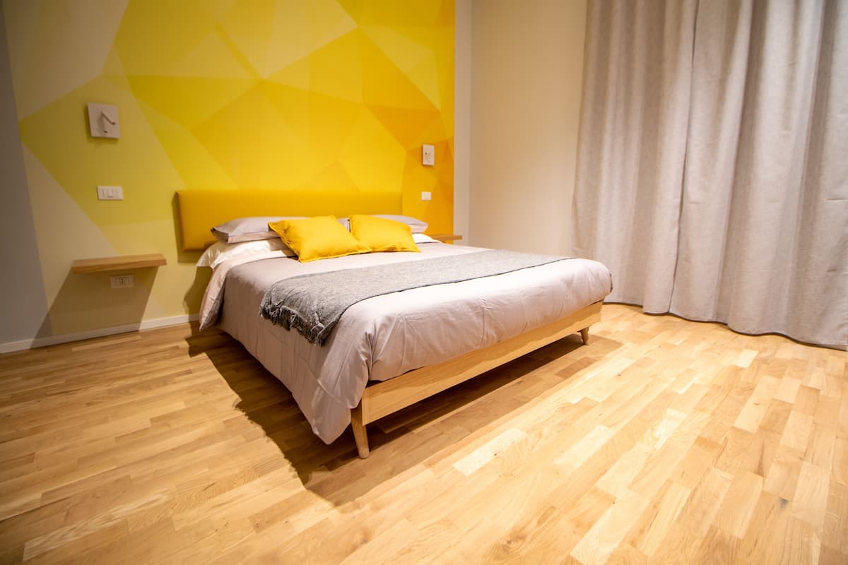 A modern bedroom with a wooden floor, a bed with a white and gray duvet, and yellow pillows.