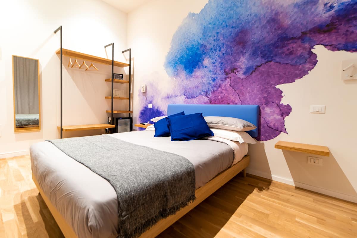 A bedroom with a bed, a wardrobe, a shelf, and a wall with a textured mural.
