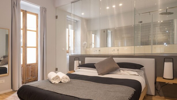 A modern bedroom with a large bed, a bedside table, a mirror, and a window with blinds. - Terrazza Sangiuliano (Catania)