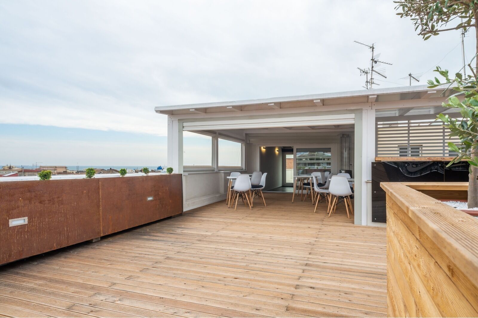 Eine Dachterrasse mit Holzboden, weißen Stühlen und Blick auf das Meer.