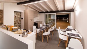 A modern kitchen with white cabinets, a wooden countertop, and a kettle. There are white chairs around a table with black placemats. A brick wall and a glass door leading to a dining area are visible. - Terrazza Sangiuliano (Catania)
