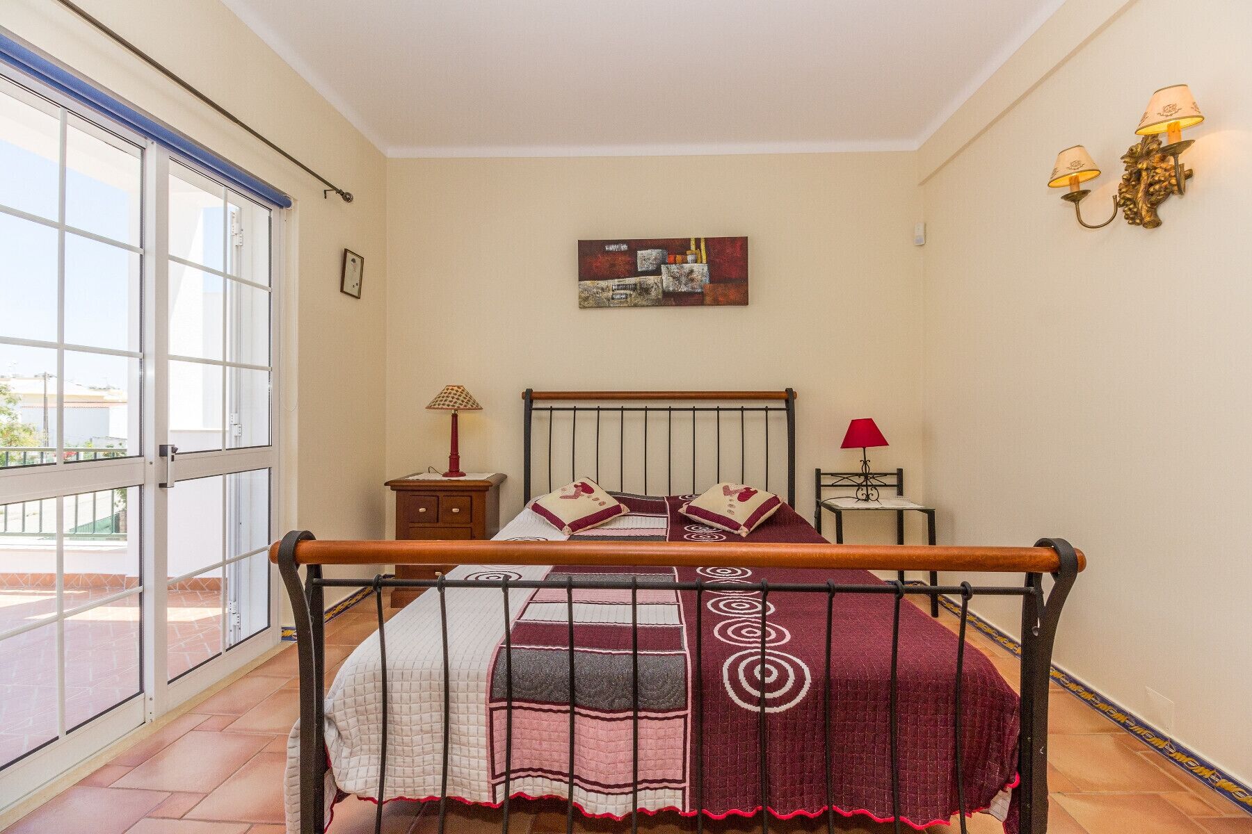 A bedroom with a bed, a nightstand, a lamp, a picture on the wall, and a view through a window.