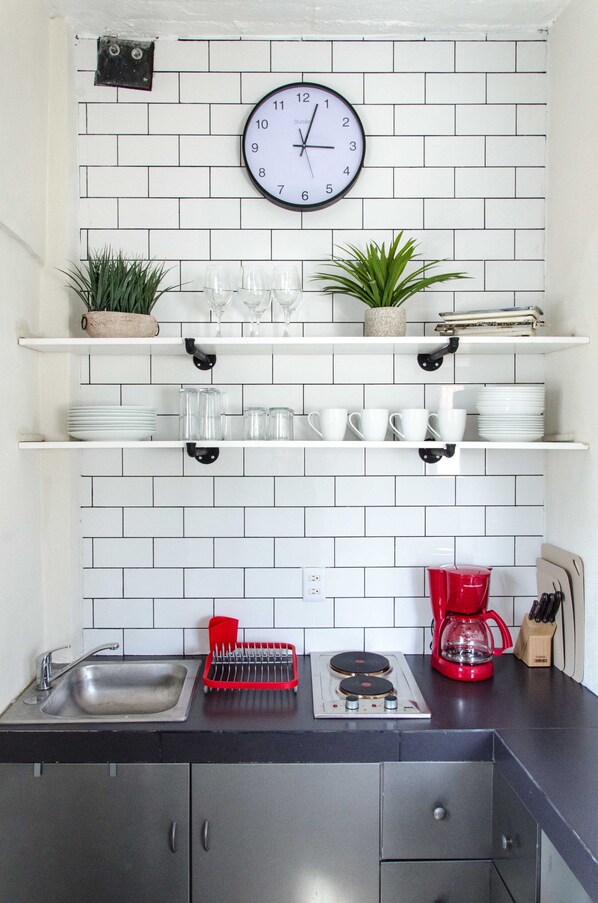 Une cuisine avec un mur recouvert de carreaux blancs, une horloge, des étagères où sont rang és des verres, un évier, une cuisinière et une machine à café.
