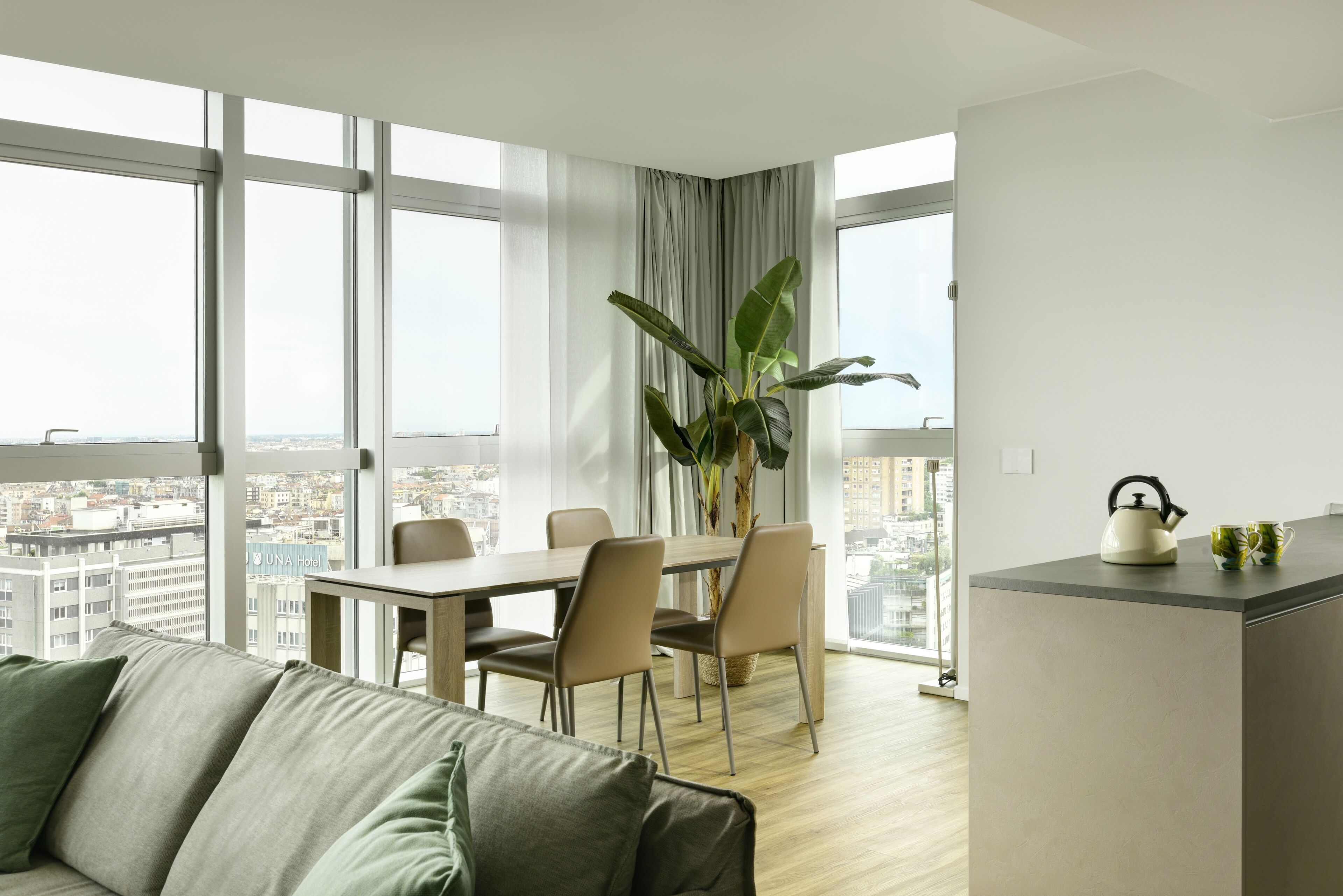 A modern living room with a sofa, a dining table with chairs, a large window with a city view, and a kitchen counter with a kettle and mug.