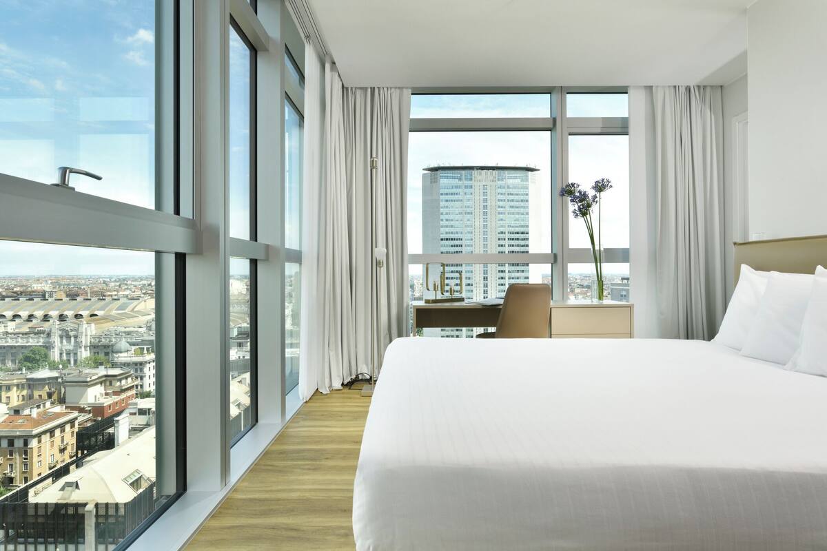 Habitación de hotel con una cama grande, un escritorio y vistas a un paisaje urbano a través de amplias ventanas.