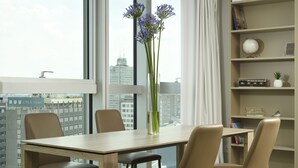 Una habitación de hotel moderna con una mesa de comedor, sillas, un jarrón con flores, una estantería y vistas a la ciudad.