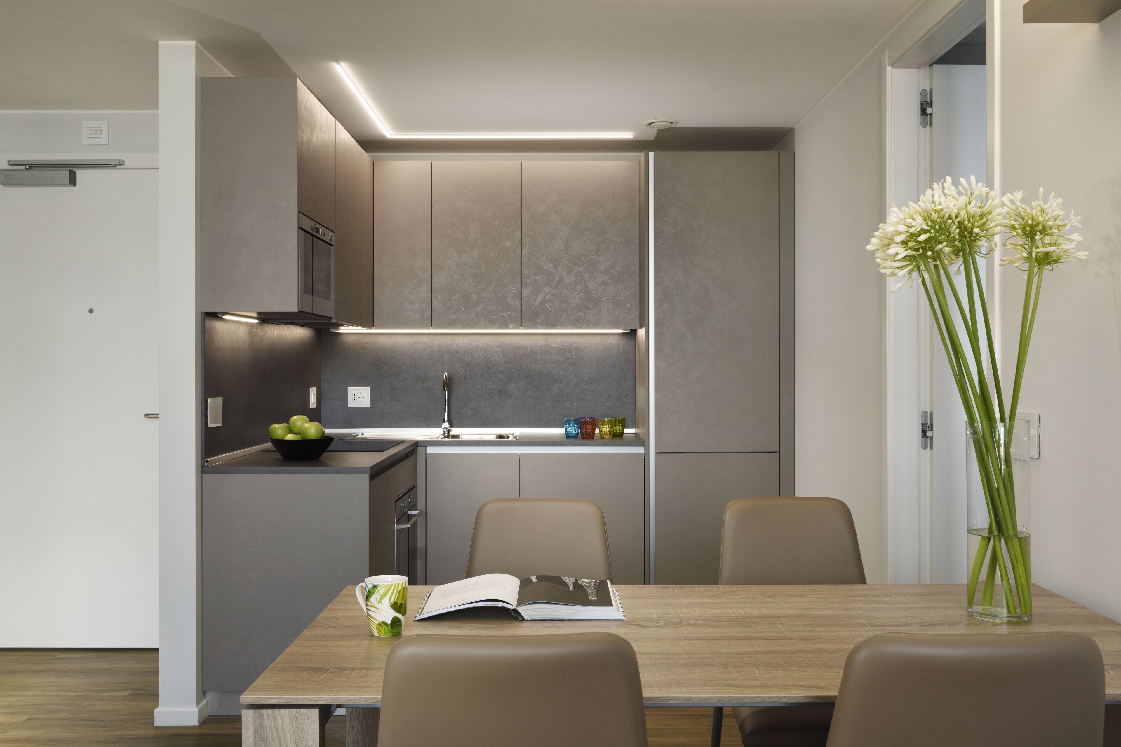 A modern kitchen with a built-in stove, a dining table with chairs, and a vase with flowers on the table.