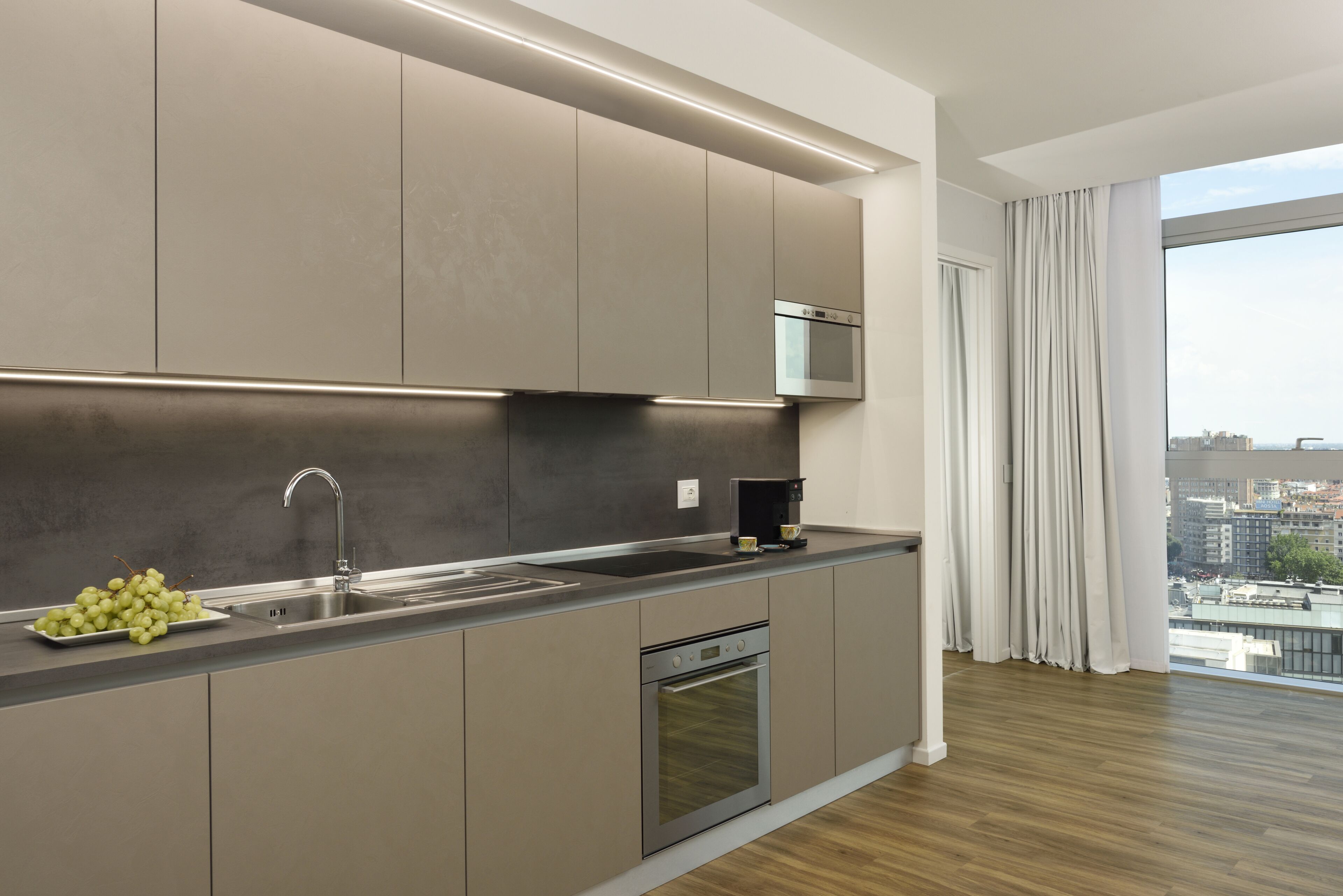 A modern kitchen with a built-in oven, stainless steel sink, and a large window offering a city view.