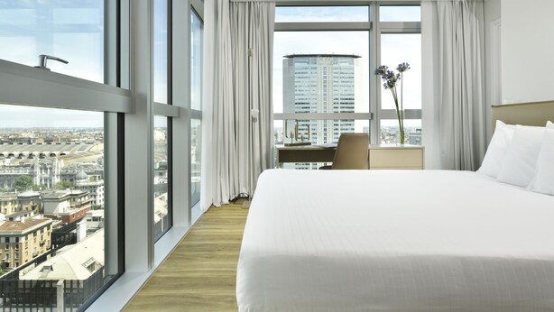 Habitación de hotel con una cama grande, un escritorio y vistas de la ciudad a través de amplios ventanales.