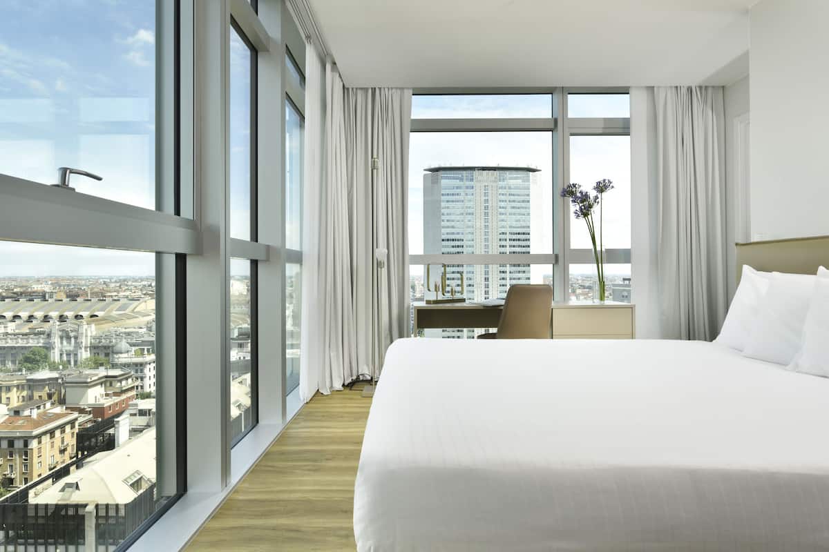 Habitación de hotel con una cama grande, un escritorio y vistas de la ciudad a través de amplios ventanales.