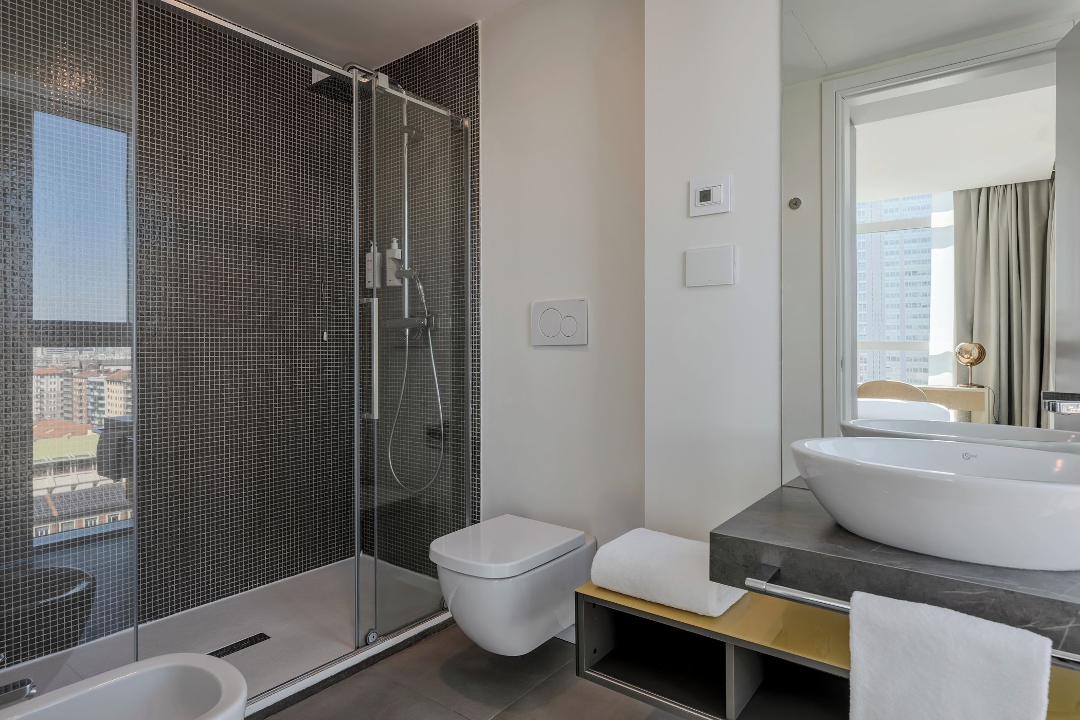Ein modernes Badezimmer mit einer gläsernen Duschkabine, einer weißen Toilette, einem großen Waschbecken und einem Spiegel, der den Blick auf die Stadt reflektiert.