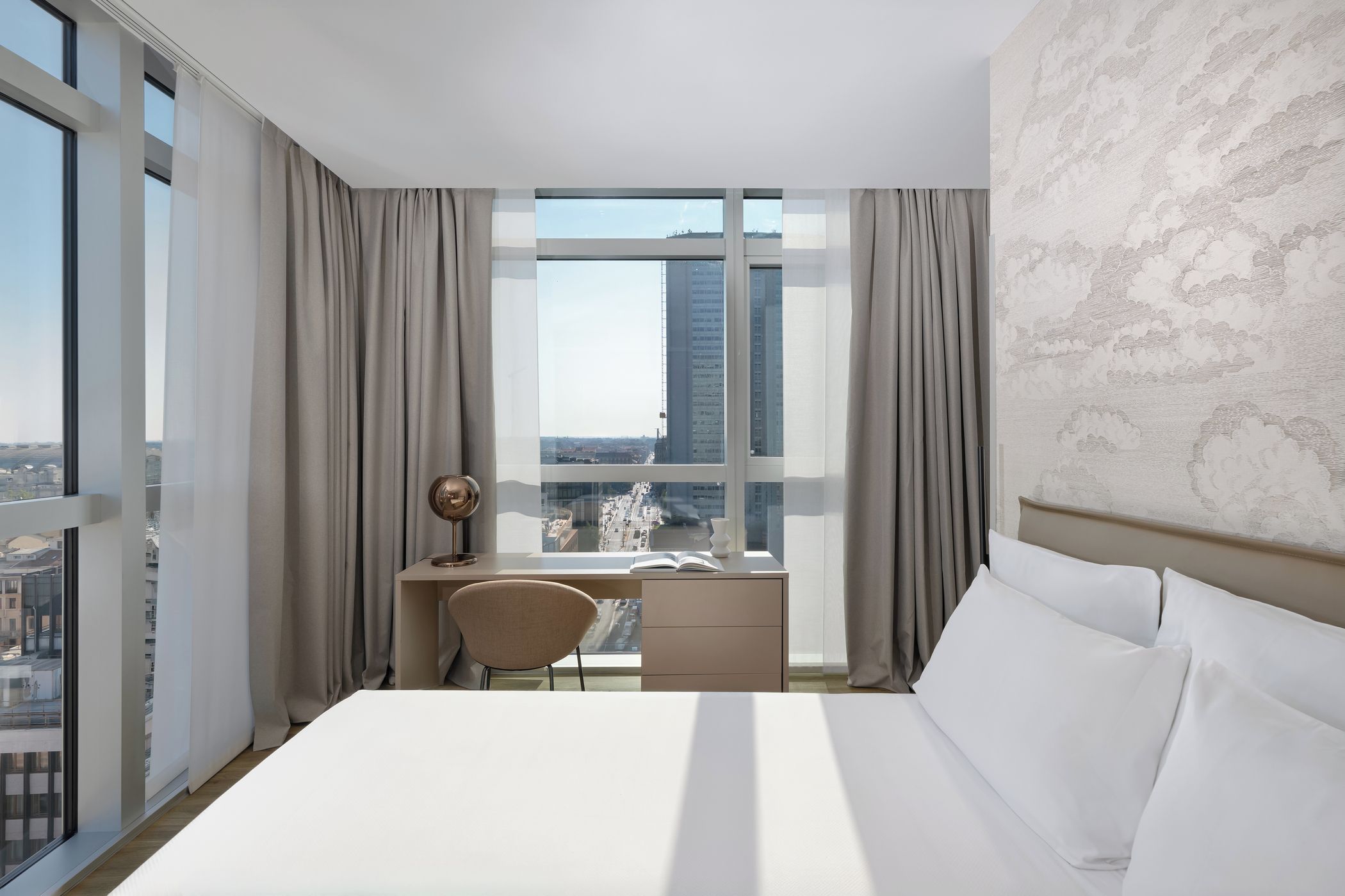 A hotel room with a bed, a desk, and a chair. There is a large window with curtains, and a view of buildings outside.