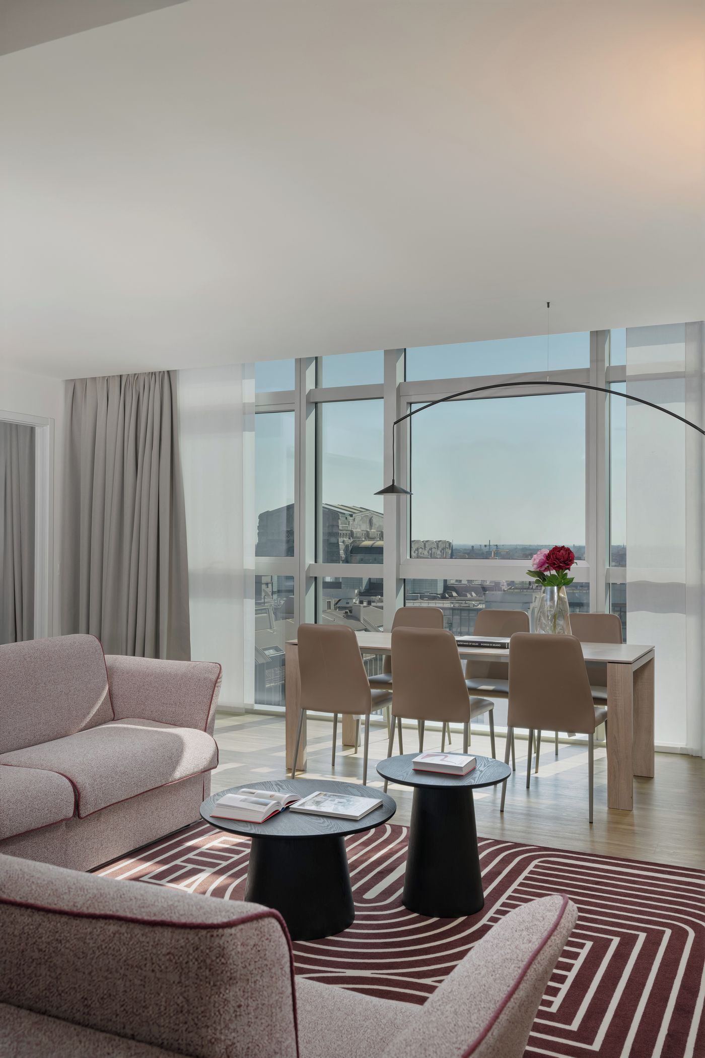A modern living room with a sofa, a dining table with chairs, a large window with a view, and a floor lamp.