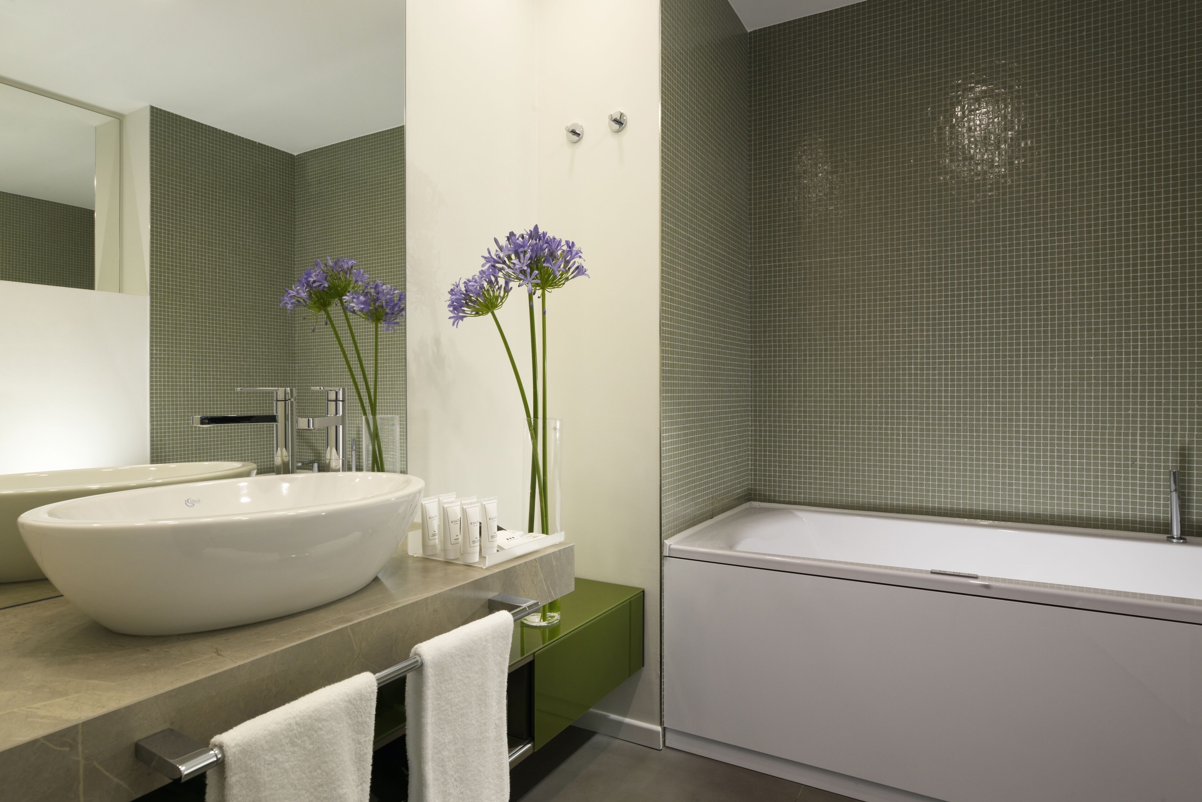 A modern bathroom with a large white sink, a green vanity, a white bathtub, and two purple flowers on the vanity.