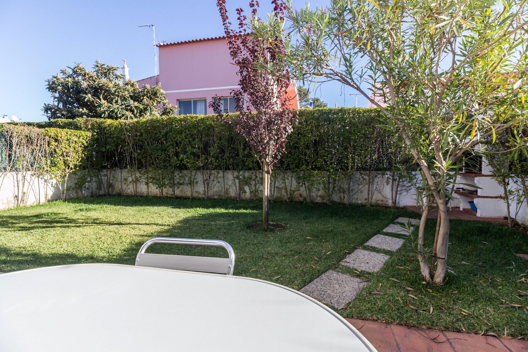 A well-maintained garden with a white table and chairs, a hedge, and a pink building in the background.