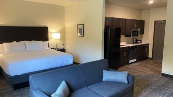 A hotel room with a bed, a grey sofa, a small kitchen area with a refrigerator and microwave, and a nightstand with a lamp.