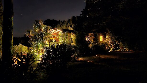 Ein Haus mit einer großen, beleuchteten Kuppelkonstruktion und einem kleineren Gebäude mit einem erleuchteten Fenster, umgeben von dichter Vegetation und einer felsigen Klippe.